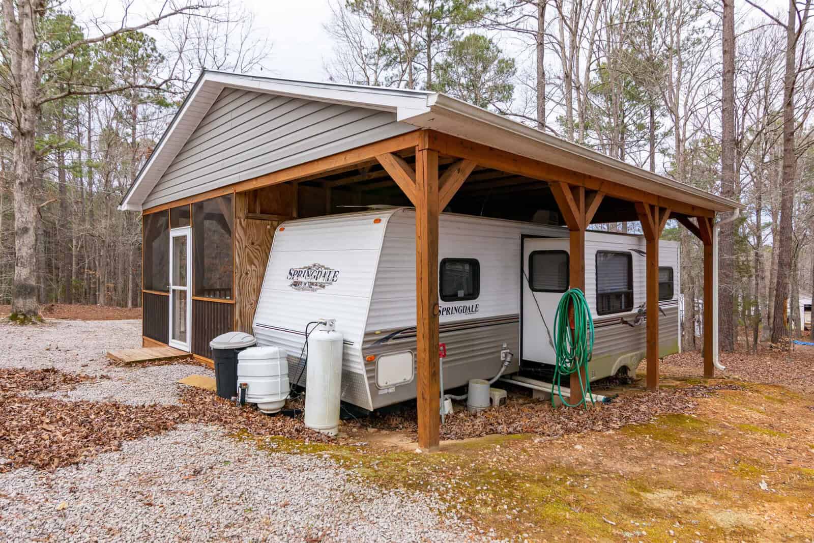 A travel trailer is parked under a wooden carport next to a screened porch in a wooded area. The trailer has hoses and propane tanks attached. The ground is covered with gravel and some patches of grass.