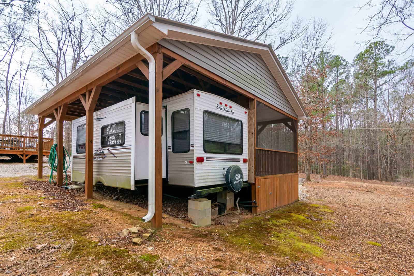 A white travel trailer is parked under a wooden carport with a gabled roof. The trailer is elevated on concrete blocks, and a green hose is coiled nearby. Trees and brown ground cover surround the area.