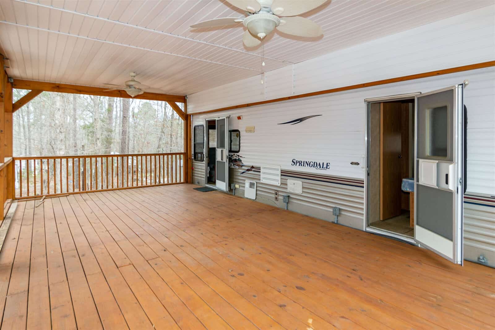 A covered wooden deck with ceiling fans is attached to a white Springdale camper. The camper’s door is open, and woods are visible beyond the deck railing.