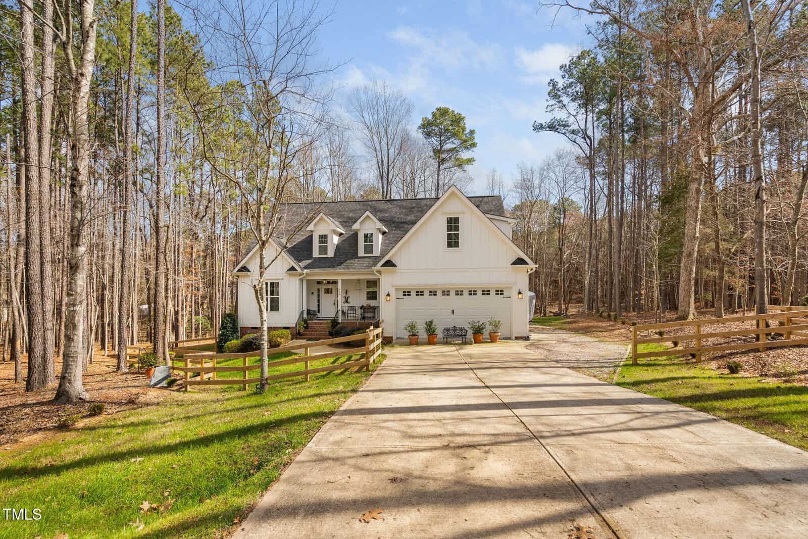 A two-story white house with a steep roof is nestled at the end of a long driveway on 564 Woodbridge Court in Wake Forest. The surrounding area is wooded, and there's a grassy lawn. A wooden fence borders the driveway, and several potted plants adorn the garage area.