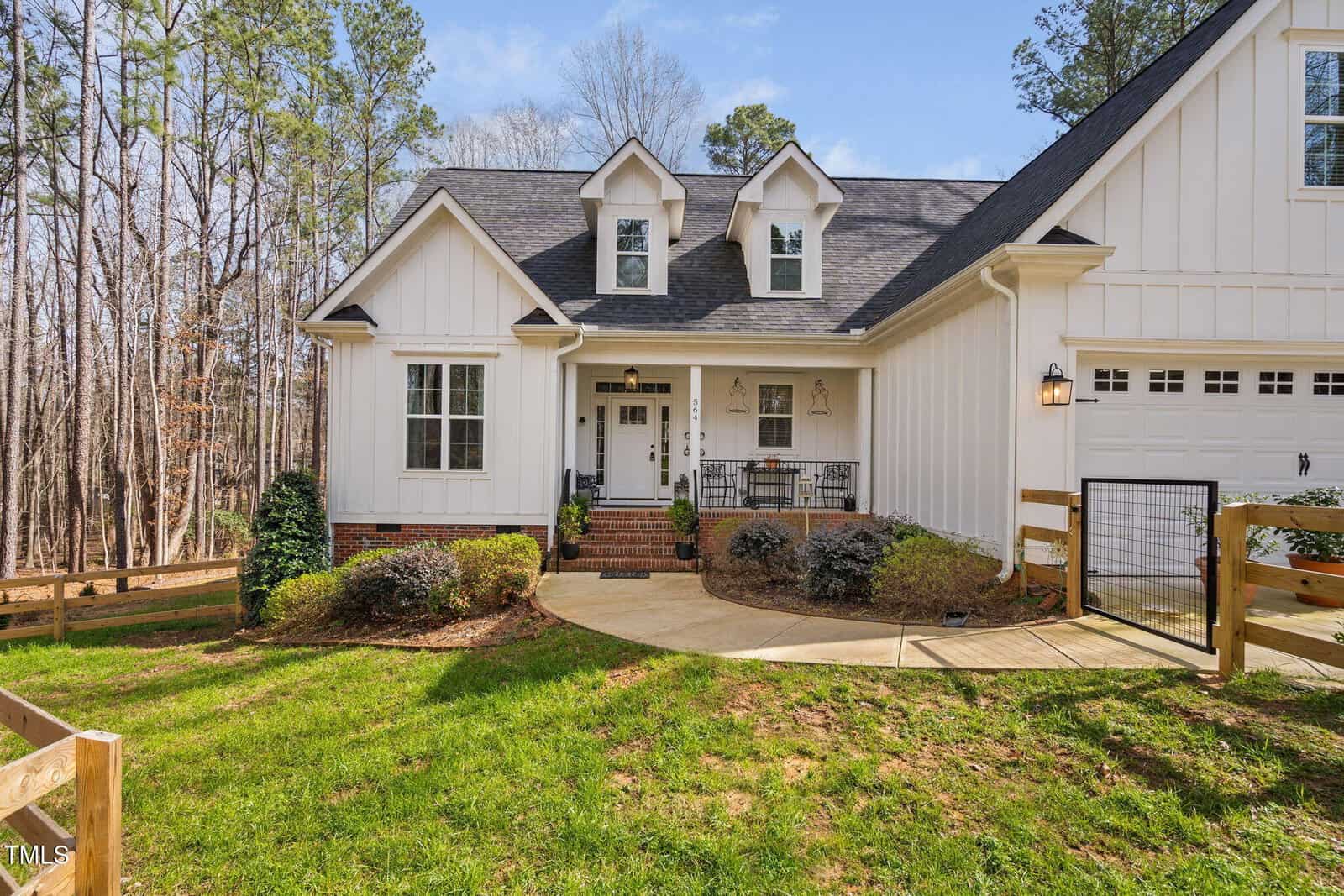 Front view of a white, two-story house with dormer windows and a porch. There's a black roof, brick steps, and a paved walkway. The house is surrounded by a grassy lawn, shrubs, and trees, with a wooden fence on the right.