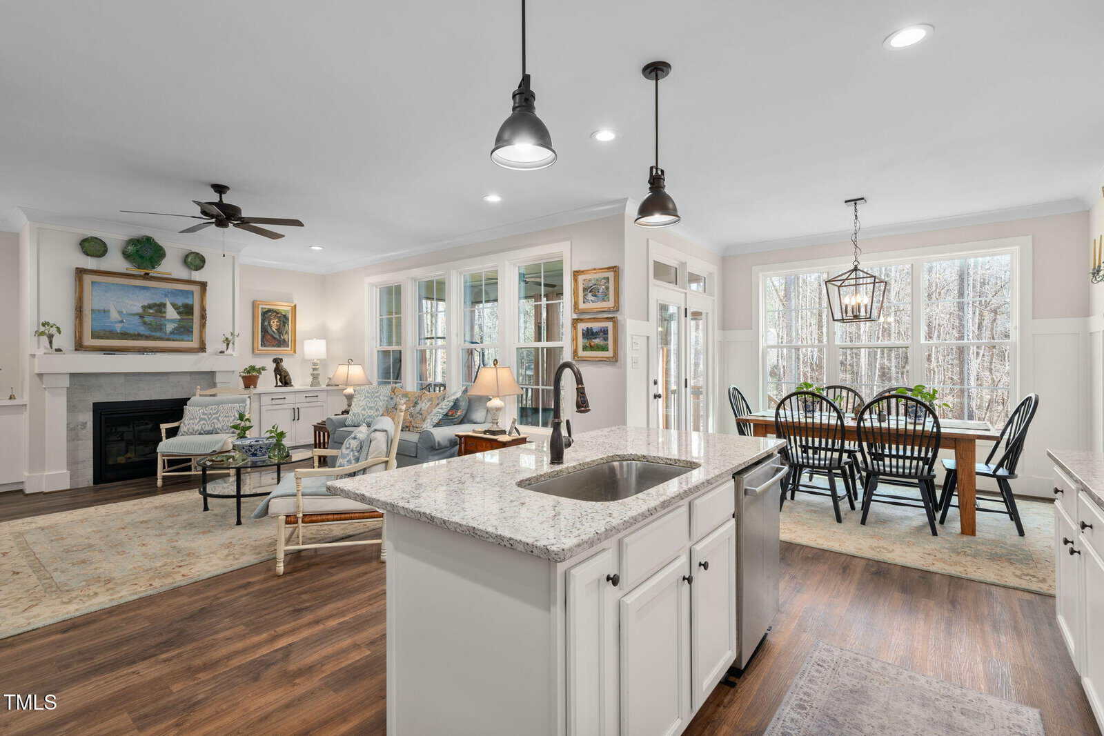 A spacious open-plan kitchen and living area with white cabinetry and a granite island. Wooden floors extend to a dining space with large windows. The living room features a fireplace, sofa, and armchairs under a ceiling fan.