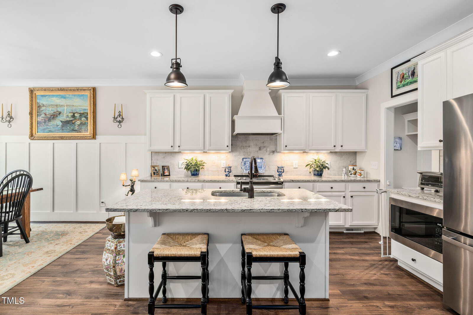A modern kitchen with white cabinets, a central island with a granite countertop, and two stools. Stainless steel appliances are present, including a refrigerator and oven. Pendant lights hang above the island, and a painting is visible on the wall.