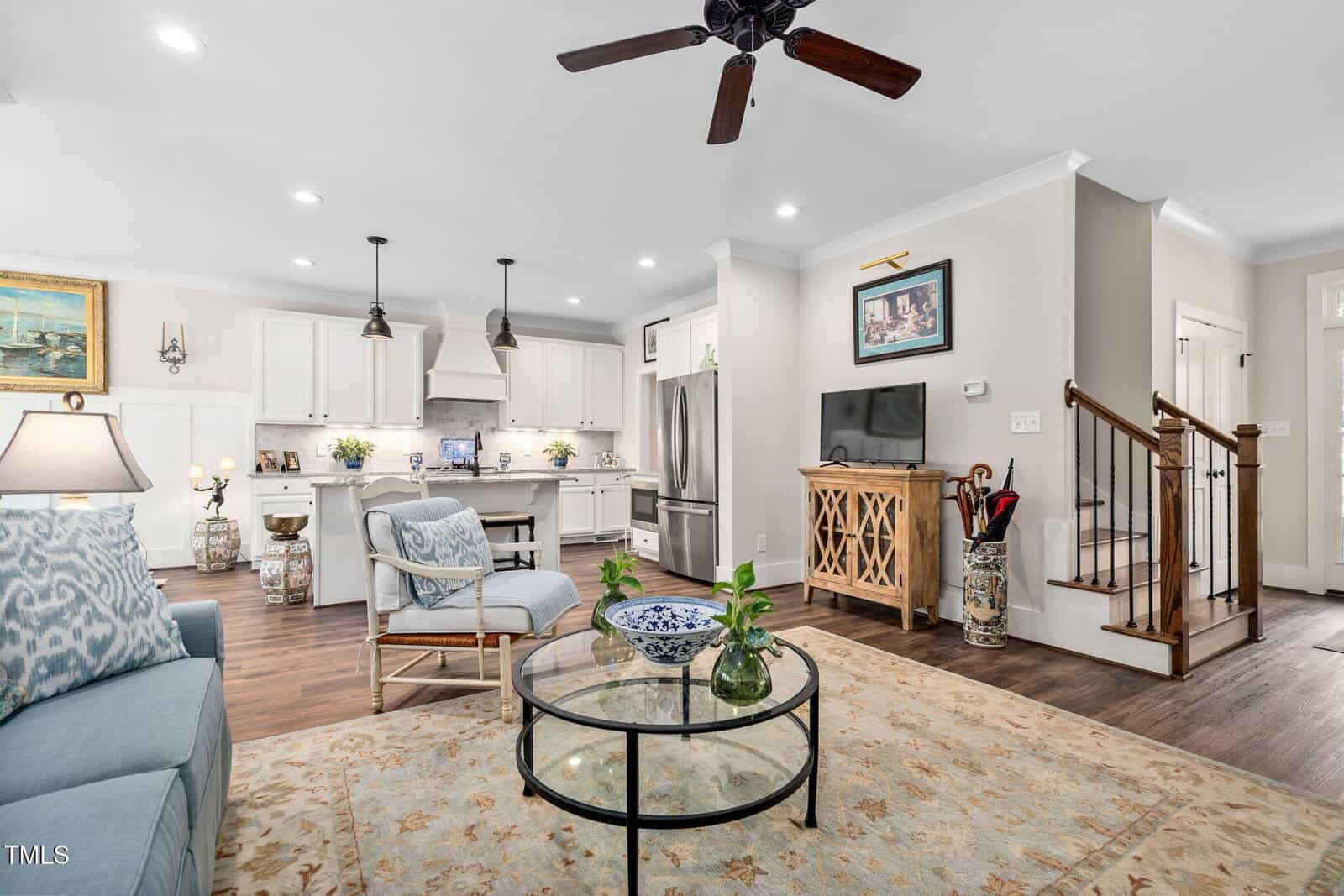 A modern living room with an open kitchen. Features include a ceiling fan, glass coffee table, light blue sofa, and white cabinets. There is a TV on a wooden stand, a decorative rug, and a staircase leading to the upper floor.