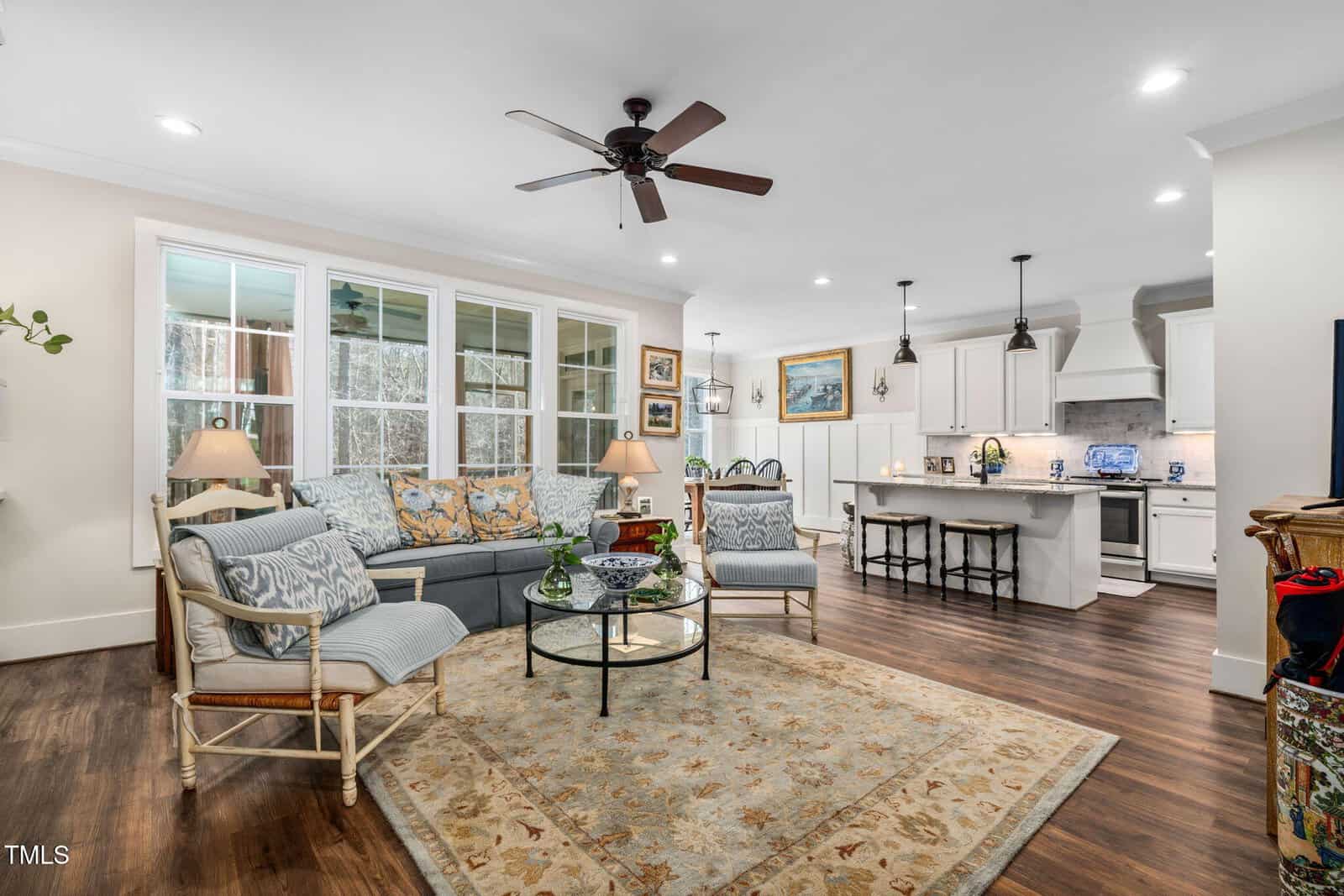 A living room with a ceiling fan above, features a light blue sofa, patterned cushions, and a glass coffee table on a decorative rug. Behind are large windows and an open kitchen with white cabinets, an island, and bar stools.