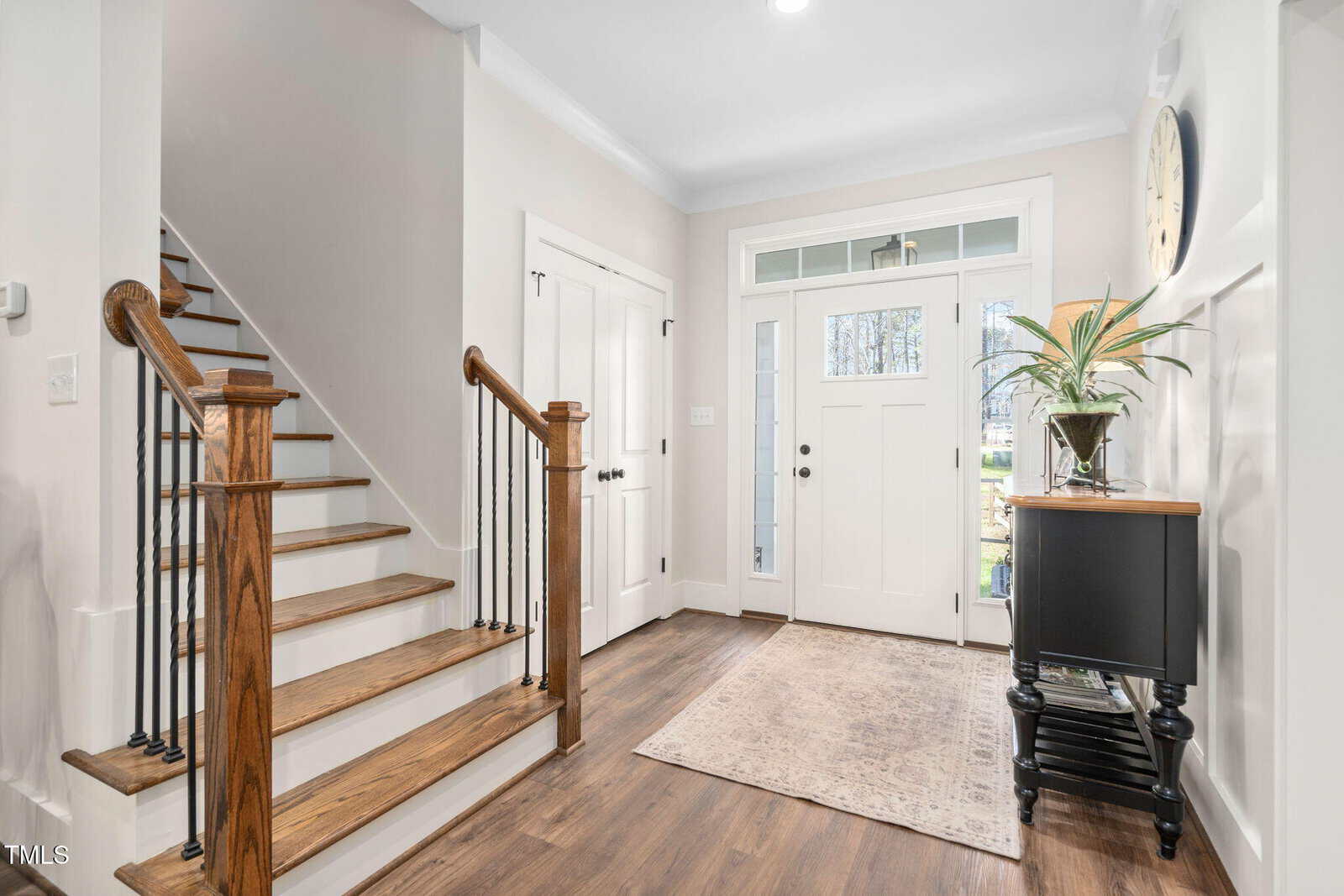 A well-lit home entryway with a wooden staircase on the left side. The floor is hardwood, and there's a rug near the front door. A small table with a plant and a clock on the wall are on the right side. The walls are painted light gray.