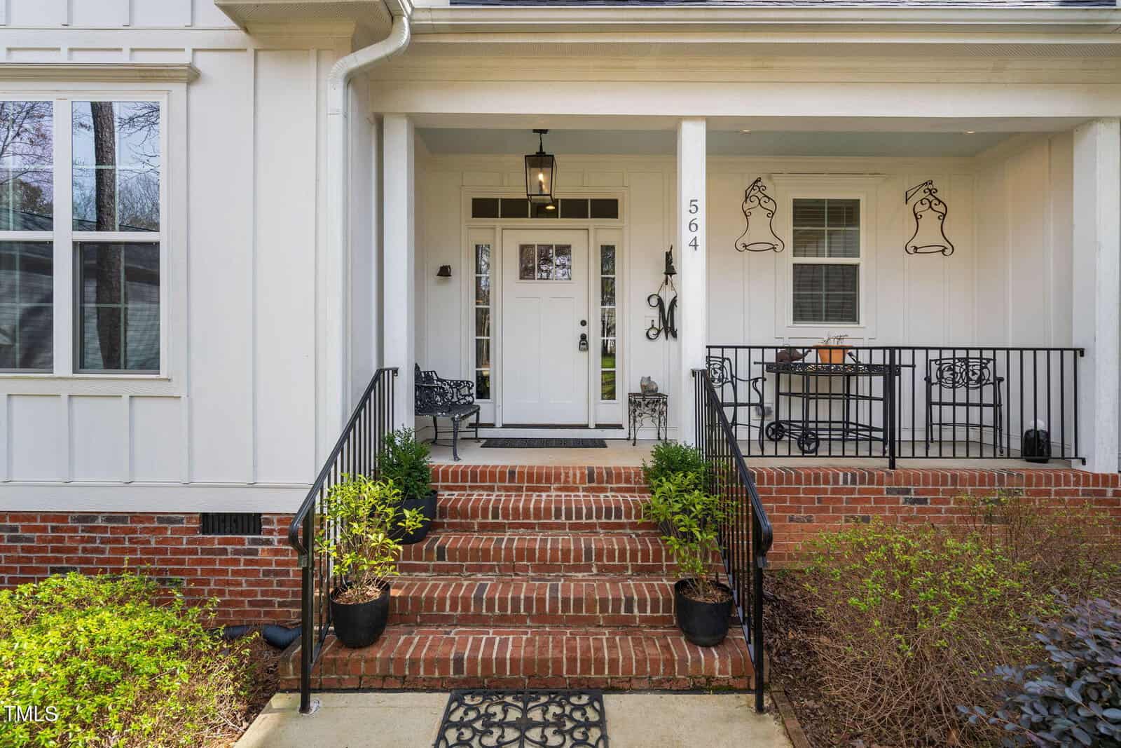 A white house entrance with a brick-lined staircase and black railings. Potted plants flank the steps. The door has sidelights and a transom window. A small bench and decorative wall art are on the porch. House number "564" is visible.