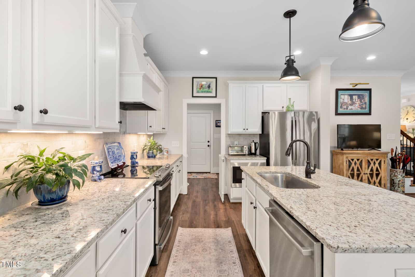 Modern kitchen with white cabinets, granite countertops, and wood flooring. Features a central island with sink, stainless steel appliances, and hanging black pendant lights. Decorative items and plants are displayed on counters and shelves.