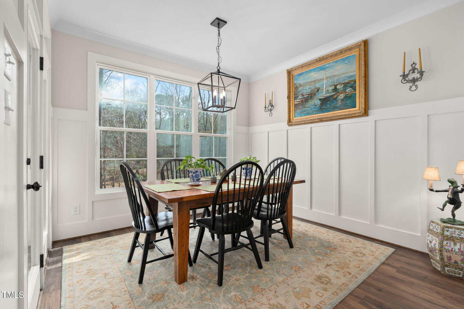 A dining room with a wooden table and six black chairs on a patterned rug. A large window with a view of trees is in the background. The walls have white paneling, and a painting and two wall sconces are above the table. A chandelier hangs from the ceiling.
