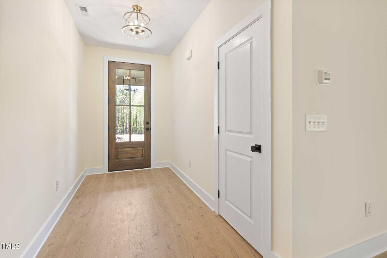 A bright hallway with light wood flooring, cream walls, a white door on the right, a thermostat, and a glass-paneled wooden front door at the end. A modern chandelier hangs from the ceiling.