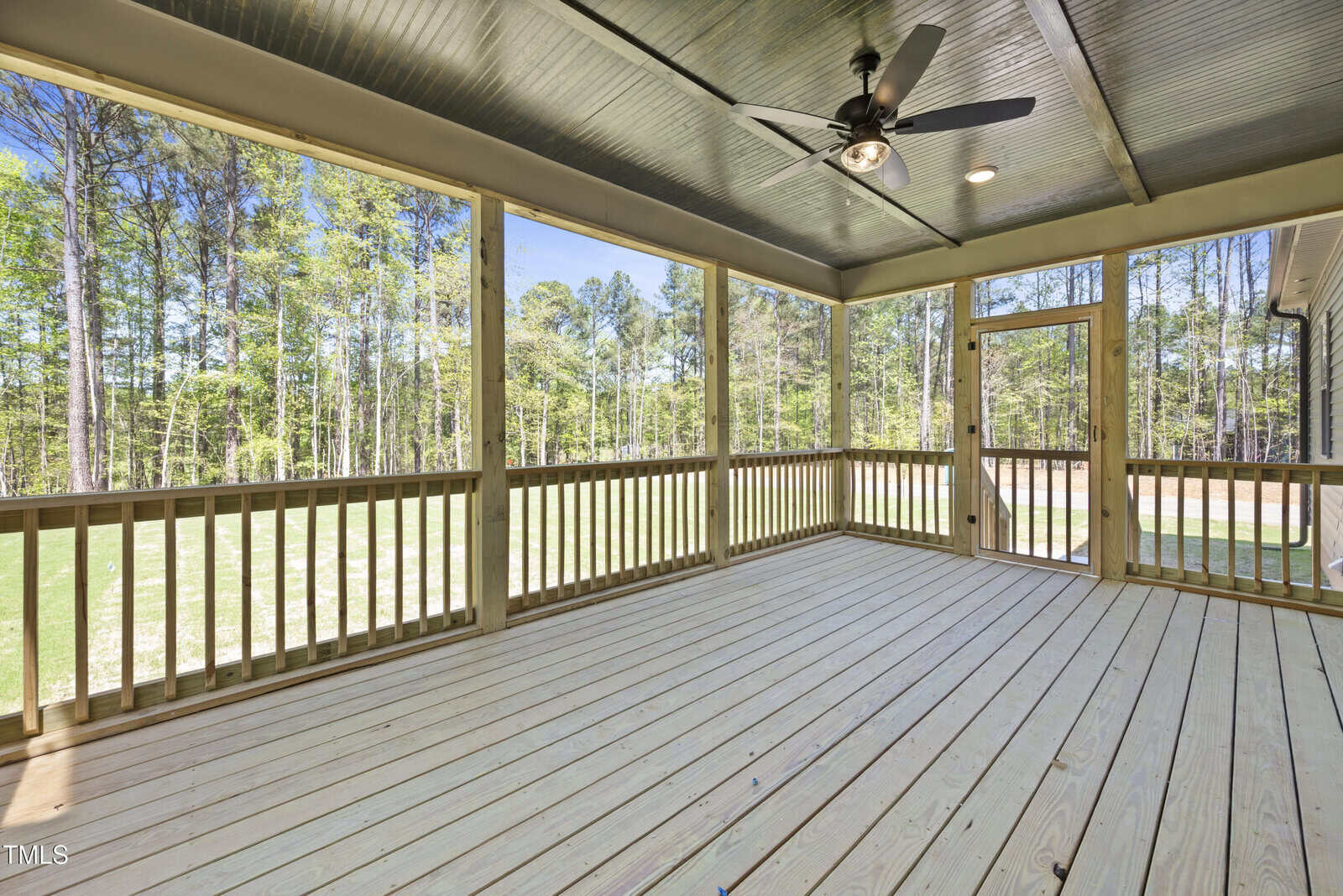 A spacious screened-in porch with wooden floors and railings, a ceiling fan, and views of a grassy backyard and tall trees beyond the screen.