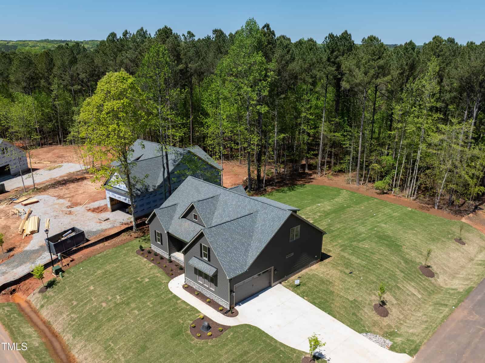 Aerial view of a modern, dark gray house with a two-car garage, surrounded by green grass and trees. A newly built home and cleared land are visible nearby under a clear blue sky.