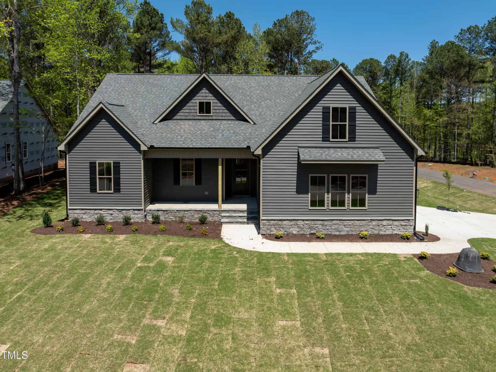 A gray two-story house with a covered front porch, gabled roof, and stone accents sits on a newly landscaped yard with green grass and a curved concrete walkway, surrounded by tall trees.