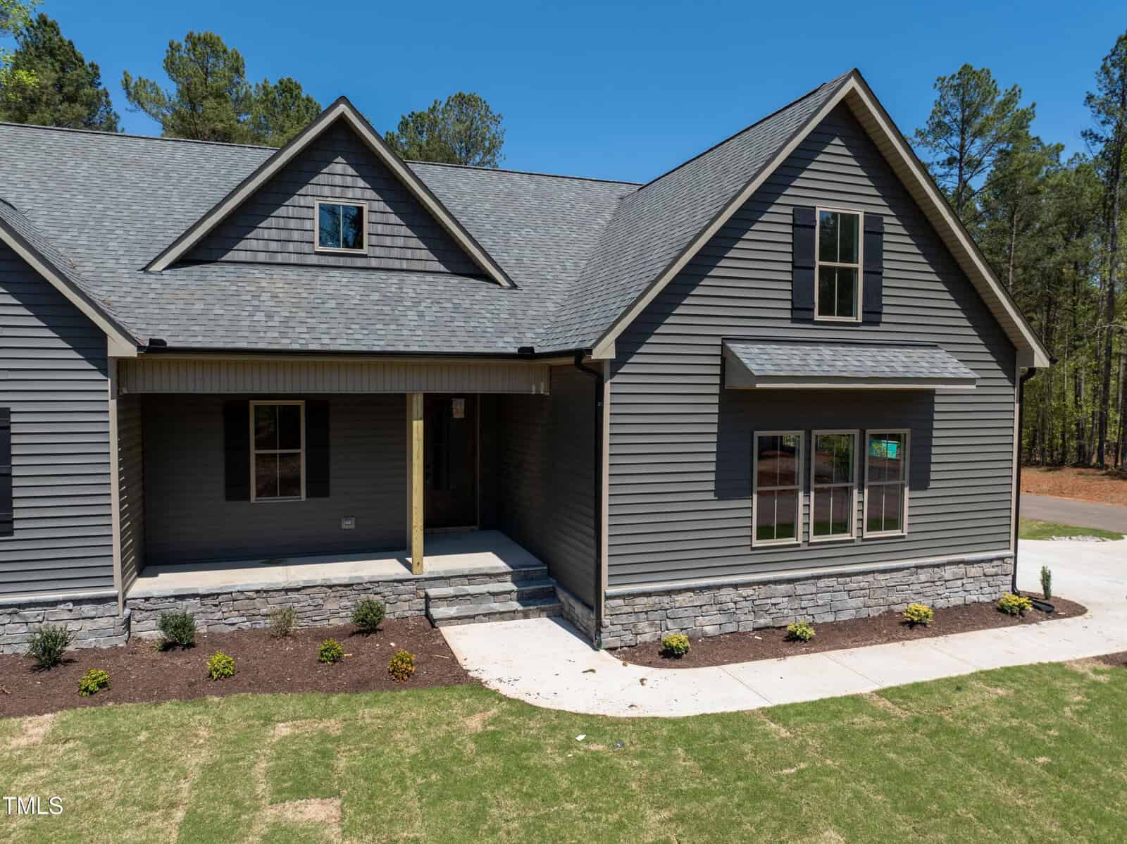 A modern gray house with stone accents, a covered front porch, and multiple windows, surrounded by a freshly landscaped yard and bordered by a concrete walkway. Tall trees are visible in the background.