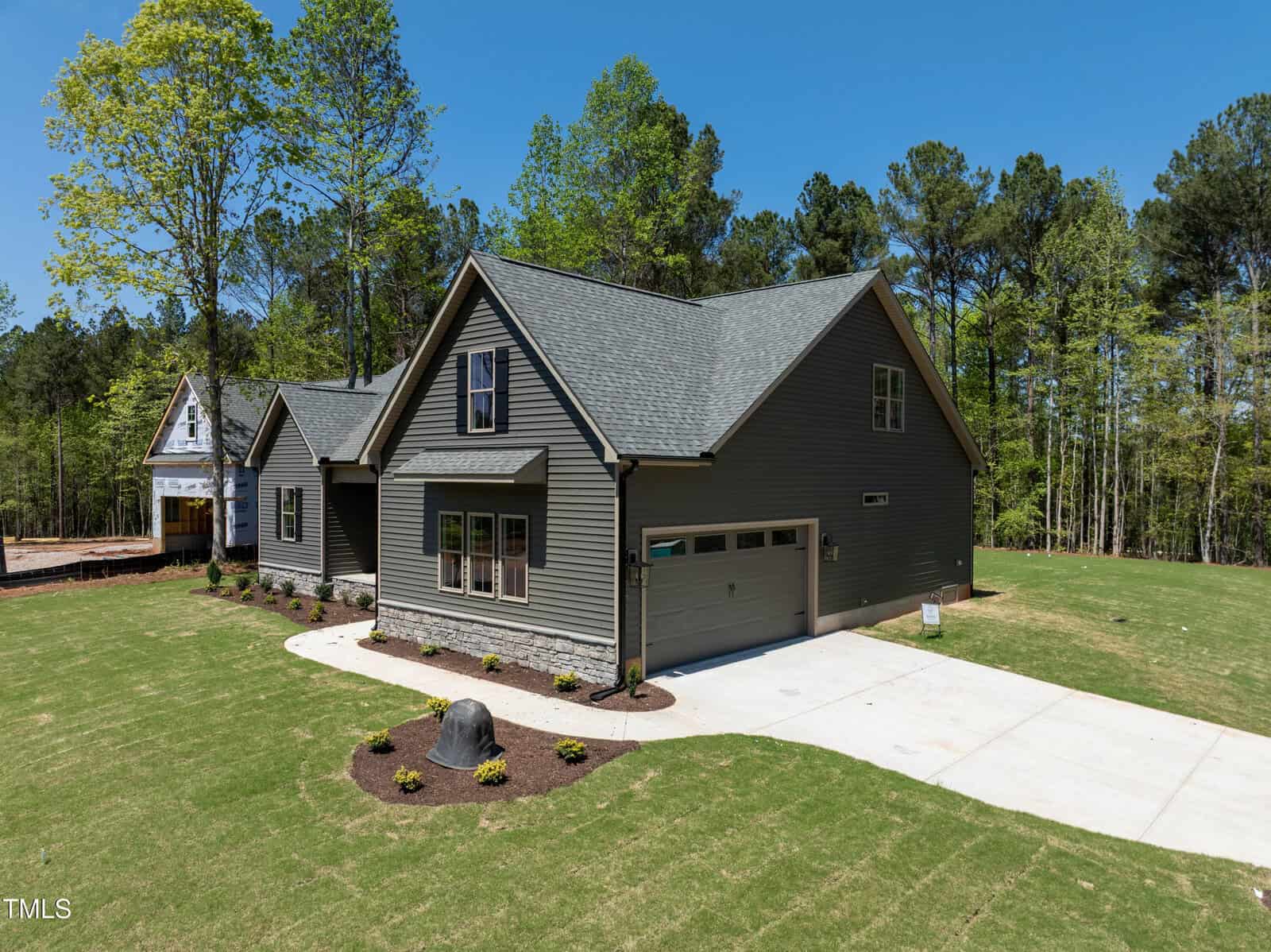 A modern two-story gray house with a double garage, neatly landscaped lawn, small shrubs, and a concrete driveway, surrounded by tall trees and another house under construction in the background.
