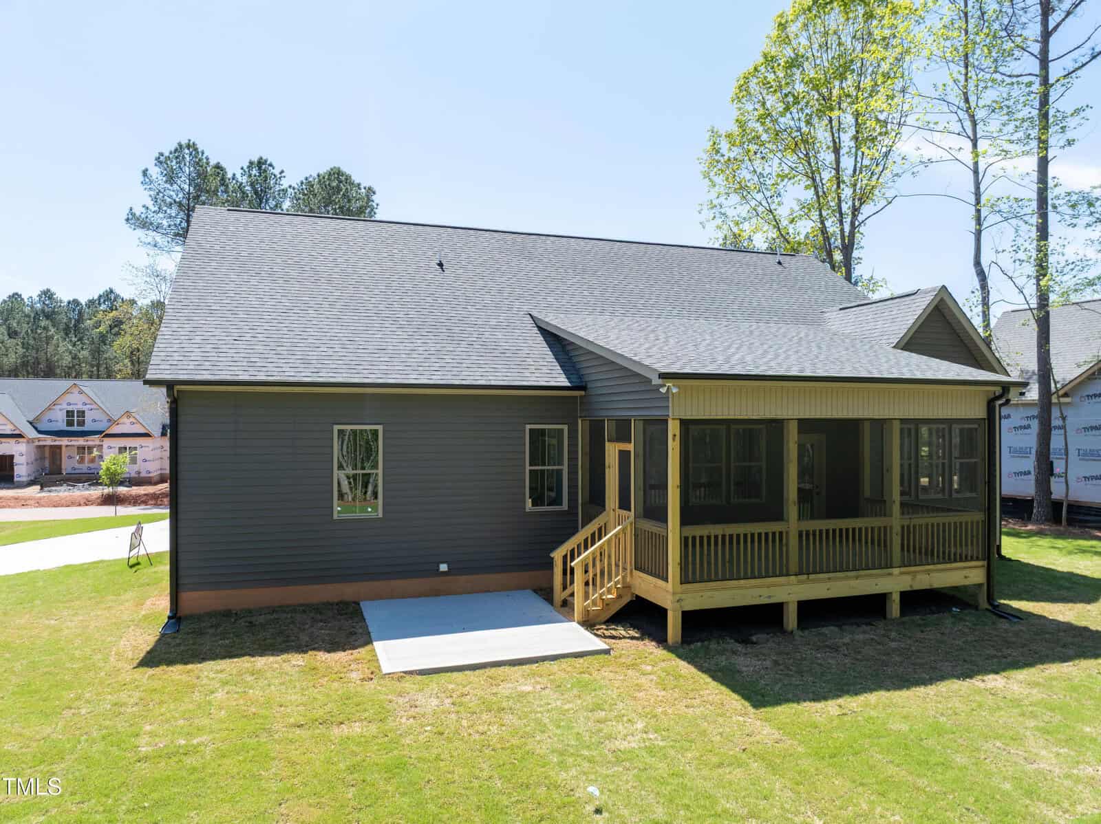 A single-story dark gray house with a gable roof, a small concrete patio, and a screened-in back porch with stairs, set on a grassy lawn on a sunny day. Other houses are visible in the background.