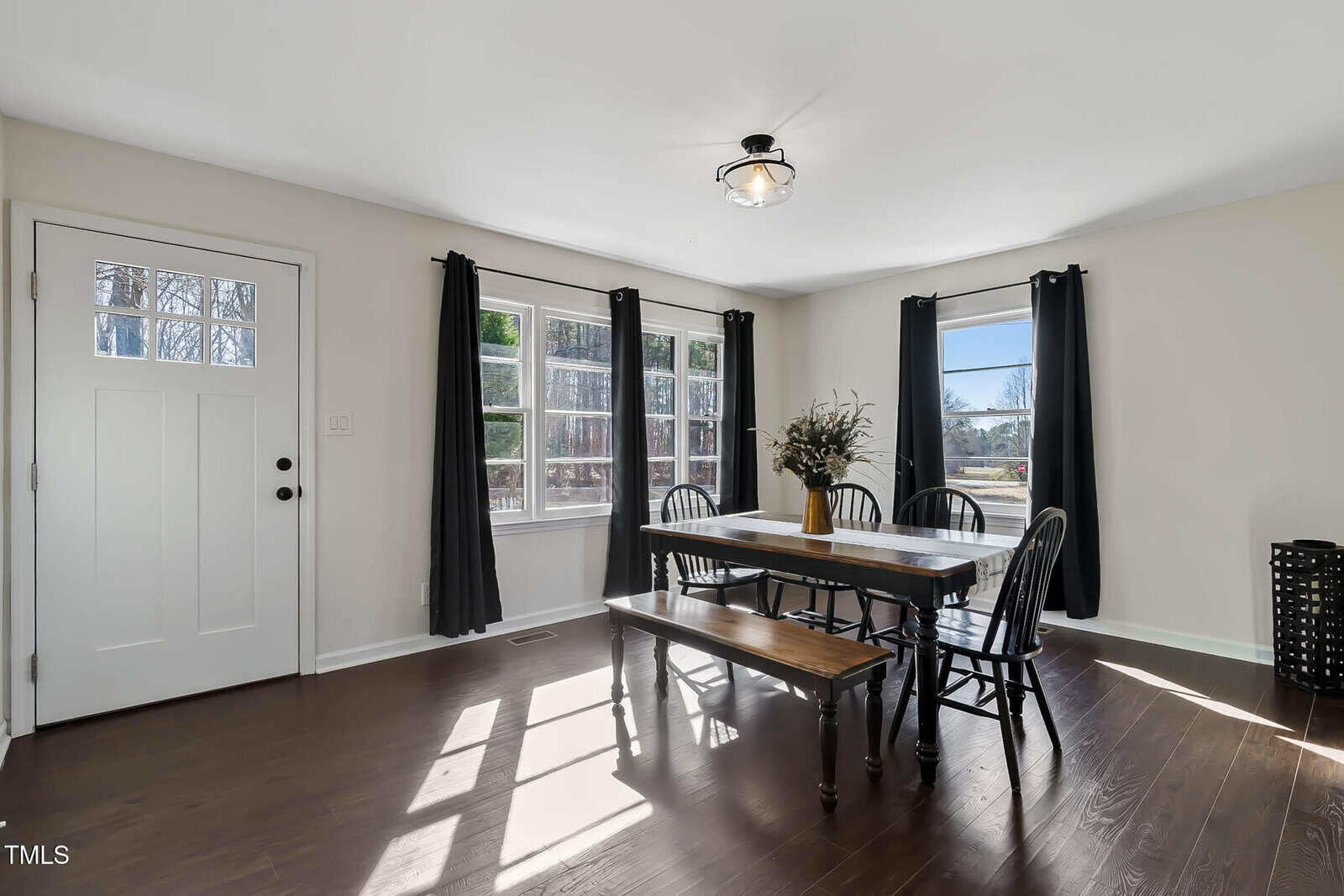 A dining room with a rectangular wooden table, six black chairs, and a wooden bench. A potted plant is on the table. Two windows with dark curtains allow natural light. A white door is on the left and hardwood floors cover the room.