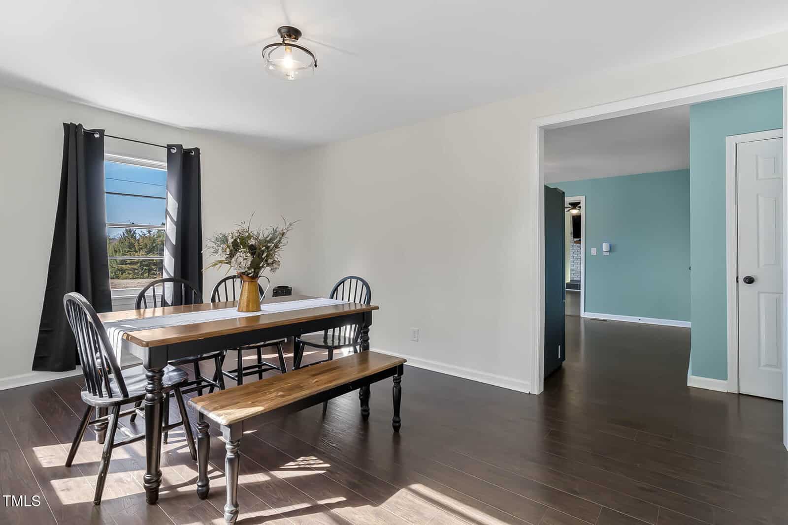 Dining area with a wooden table and benches in a room with white walls and dark wood flooring. Black chairs surround the table, which has a vase with foliage. Large window with black curtains lets in natural light. Adjacent room features a blue wall.