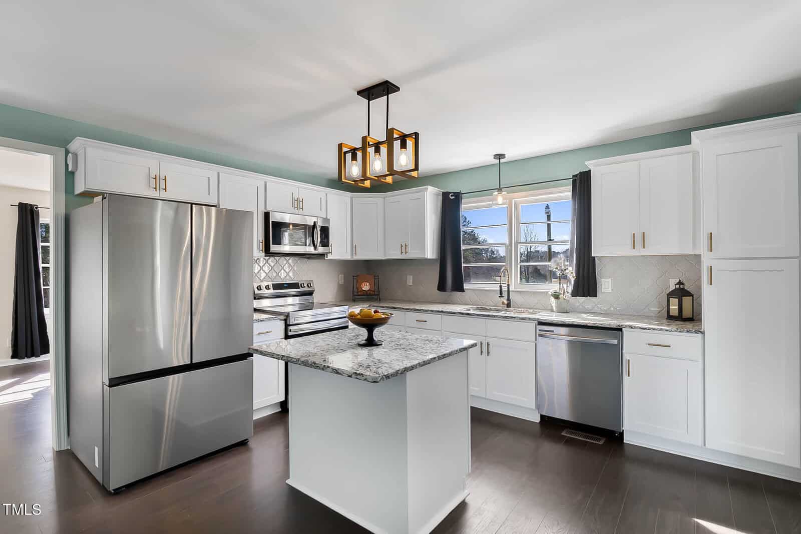 Modern kitchen with white cabinets, granite countertops, and stainless steel appliances, including a refrigerator, oven, and dishwasher. An island in the center holds a fruit bowl. A pendant light hangs above, and curtains frame the window.