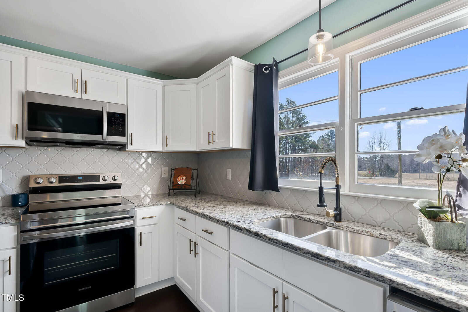 A modern kitchen with white cabinets, a stainless steel oven, and a microwave. There is a double sink under a large window with blue curtains. The backsplash is a textured tile pattern, and a potted plant sits on the counter.