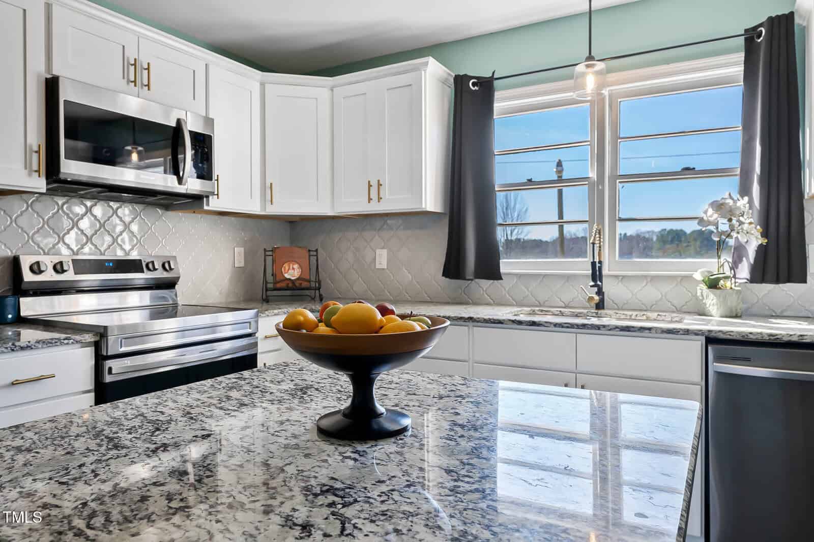 Modern kitchen with white cabinets, stainless steel appliances, and a black countertop. A fruit bowl is on the island, and a window with gray curtains allows natural light. The backsplash has a diamond pattern, and a small plant is on the counter.