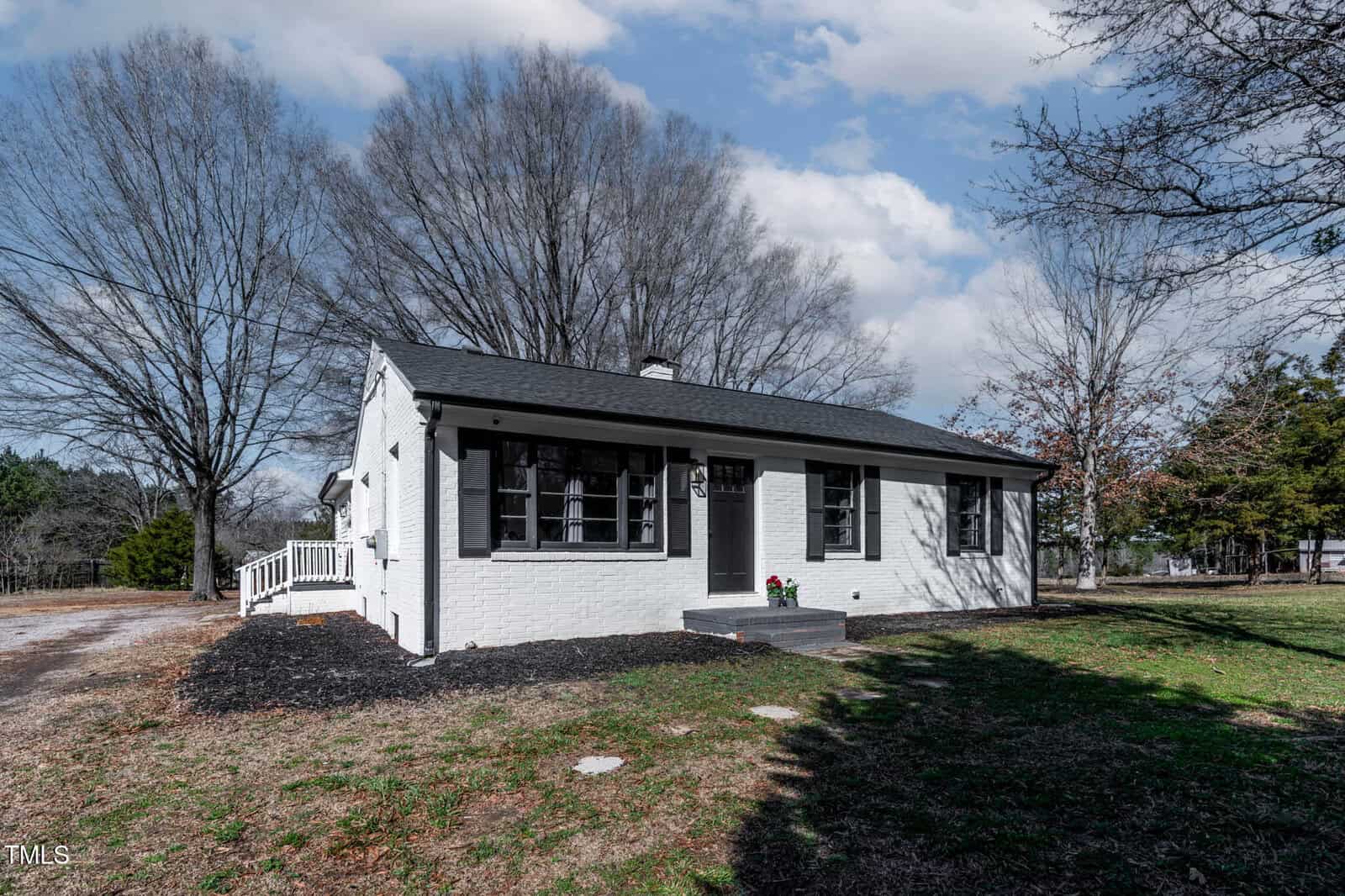 A small white brick house with a black roof and black shutters sits in a yard with sparse grass. A few large leafless trees are in the background. The sky is partly cloudy.