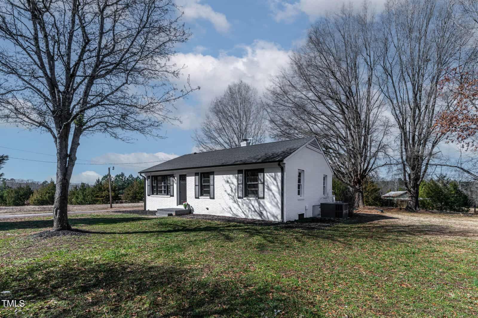 Nestled on 55 Daniel Harris Road in Henderson, this single-story white brick house features a dark roof and black shutters. It sits in an open grassy yard with a small porch, surrounded by leafless trees beneath the partly cloudy sky.