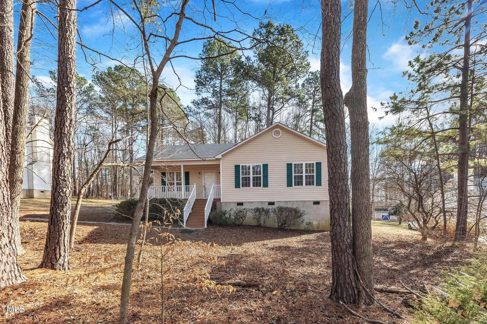 A beige single-story house with green shutters and a front porch sits among tall pine trees and leaf-covered ground on a clear, sunny day. The house has a sloped roof and a set of steps leading to the entrance.