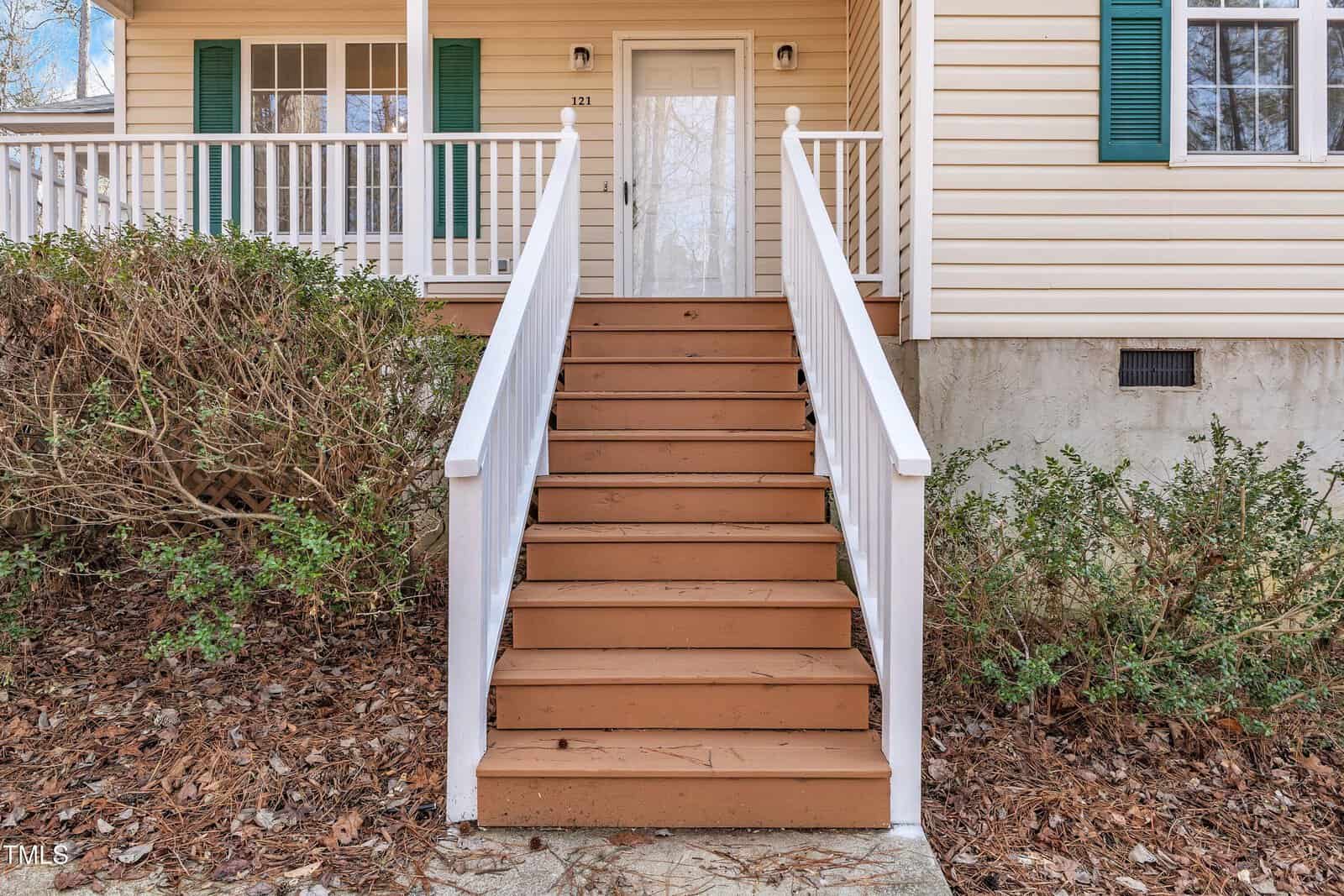 A set of brown wooden steps with white railings leads up to a white front door of a beige house with green shutters. Bushes and dry leaves are visible on either side of the steps.