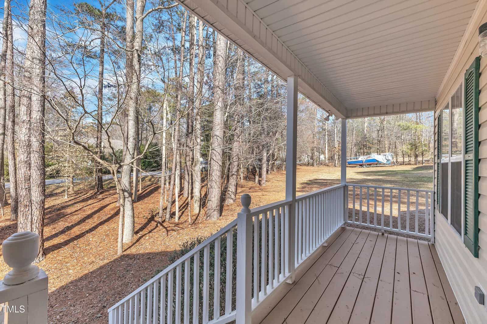 A covered wooden porch with white railings overlooks a yard with tall trees and a boat parked in the distance on a clear day.