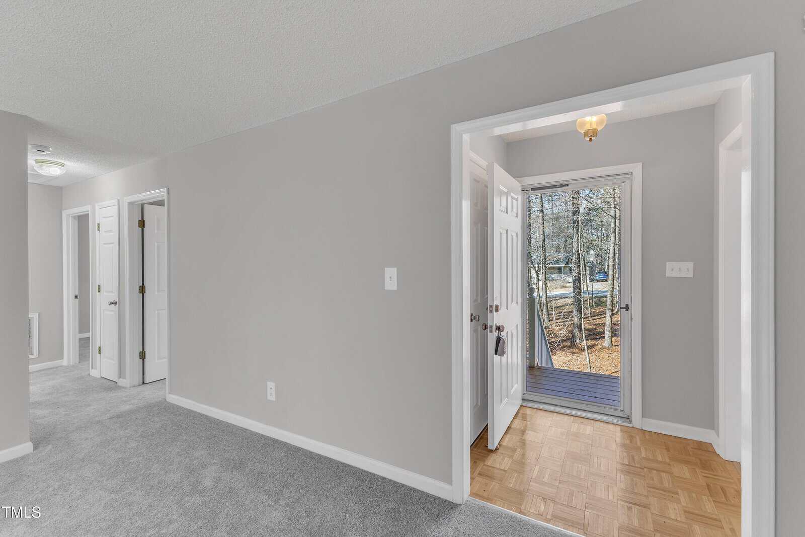 A view of a home's interior shows a carpeted hallway with several doors on the left, and a small entryway with wood flooring and a glass door leading outside to a wooded area. Walls are painted light gray.