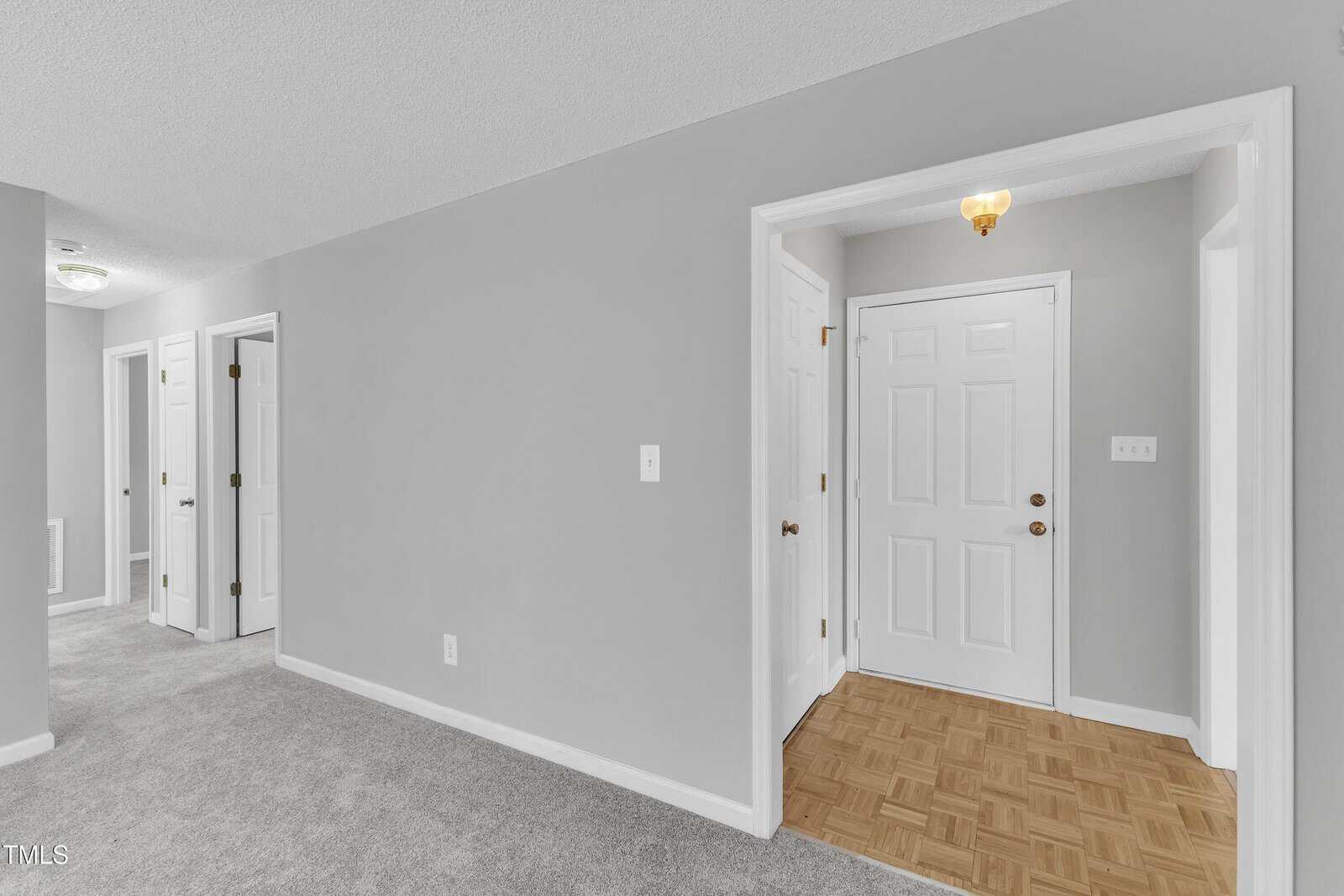 View of a hallway and entry area in a home, featuring gray walls, white doors, carpeted flooring in the hallway, and parquet flooring at the entrance. Ceiling lights are visible, and the space appears clean and empty.