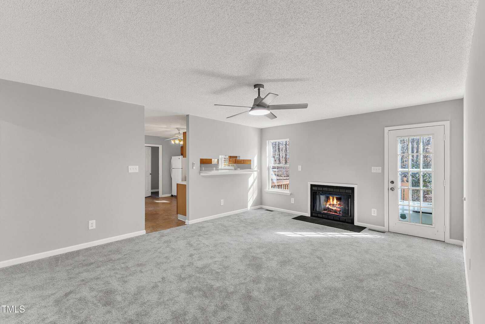A spacious living room with light gray carpet, white walls, a ceiling fan, a lit fireplace, and a glass door leading outside. The room connects to a kitchen area visible through an open doorway.