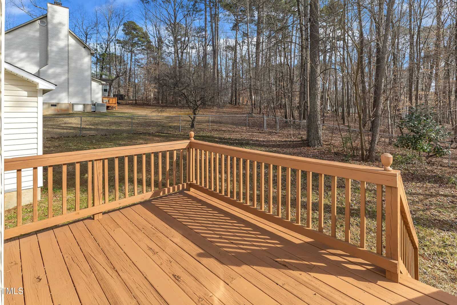 A wooden deck with railing overlooks a backyard bordered by trees and a chain-link fence. Sunlight casts shadows on the deck, and a neighboring house is visible to the left.