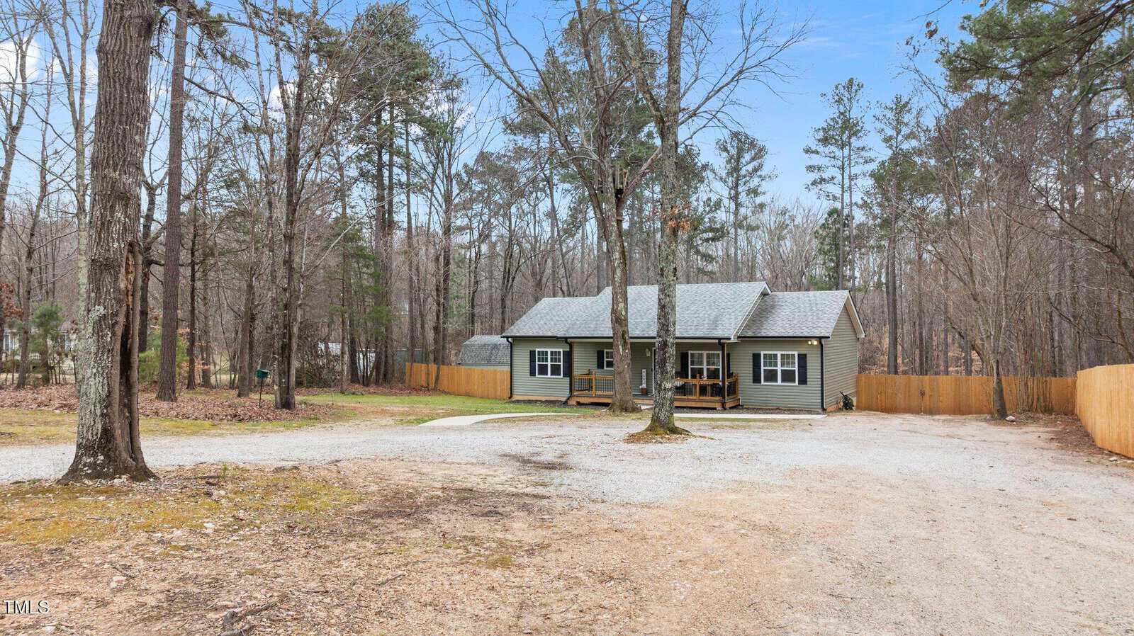A single-story house with gray siding sits in a wooded area. There is a gravel driveway leading to the house, surrounded by trees and a wooden fence. The sky is clear and blue.