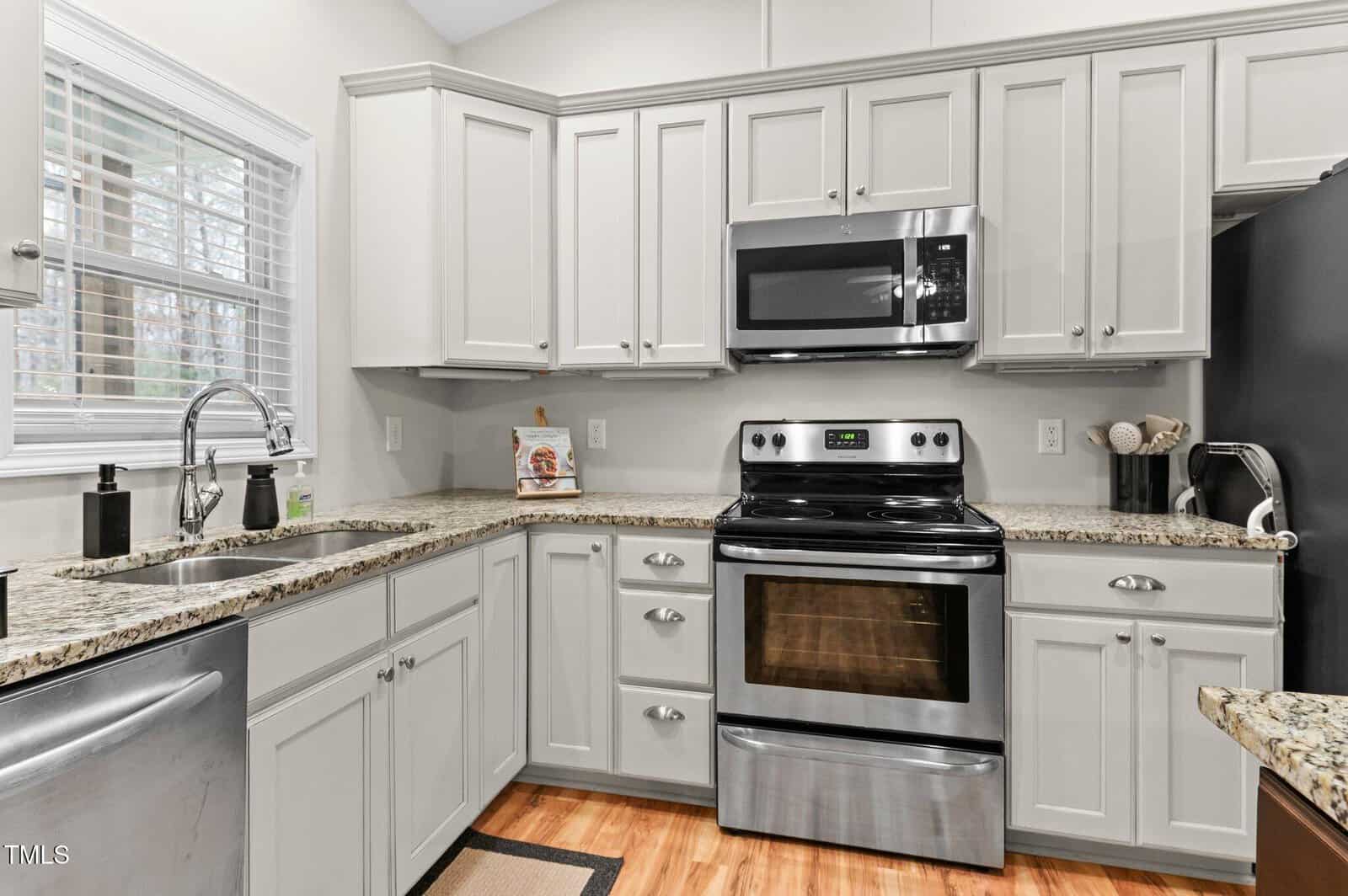 A modern kitchen with white cabinets, stainless steel appliances including a dishwasher, oven, and microwave, and granite countertops. A window with blinds is above the sink, and wooden flooring completes the space.