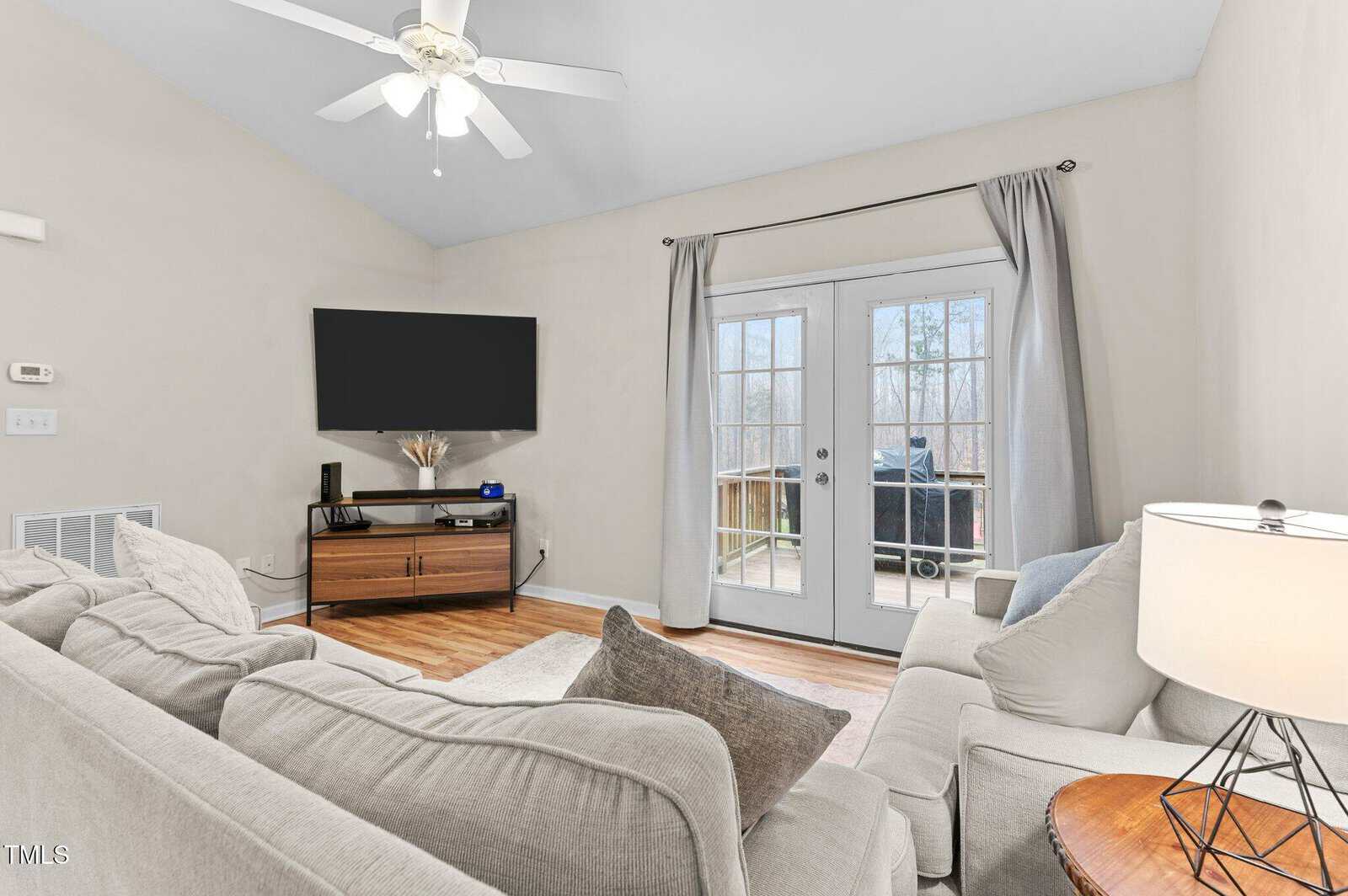 A living room with beige walls and hardwood flooring features a ceiling fan, a wall-mounted TV, and a wooden TV stand. A light gray sofa and armchair are set around a wooden side table with a lamp. French doors lead to an outdoor deck.