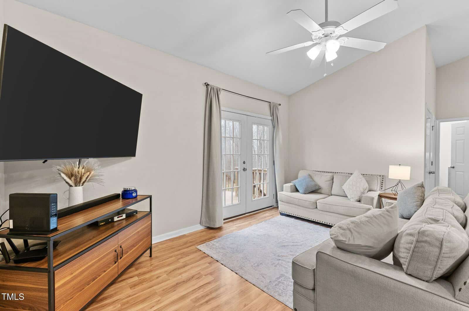 Living room with beige walls and a wooden floor. Features a ceiling fan, gray sofas, a blue area rug, and a wooden console with a large wall-mounted TV. Glass doors with curtains lead to an outdoor view. A lamp and decorative items are present.