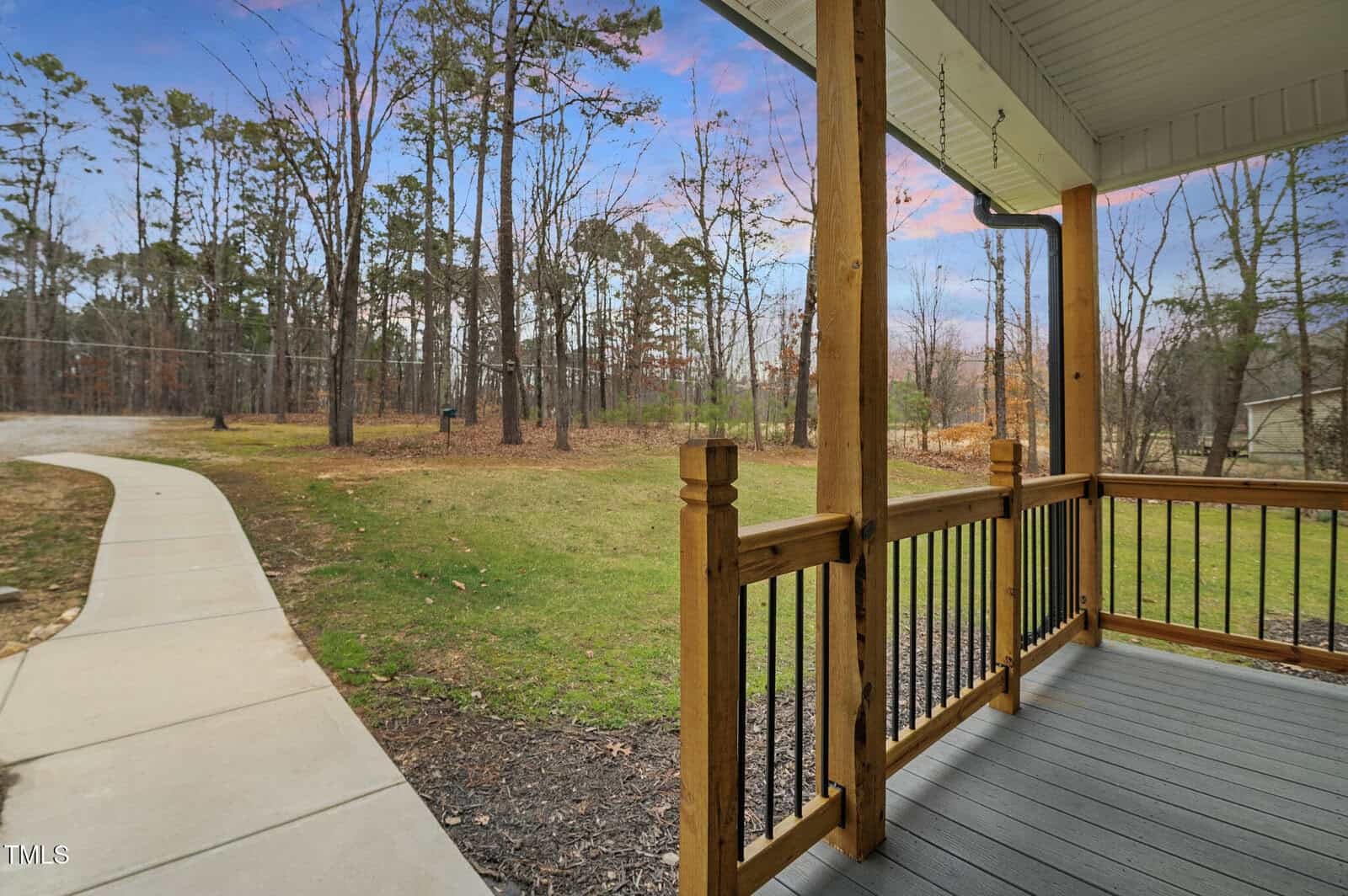 A wooden porch with black railing overlooks a yard with a paved walkway. Tall trees and a clear sky are visible in the background. The porch roof is white with decorative trim. The scene is calm and natural.