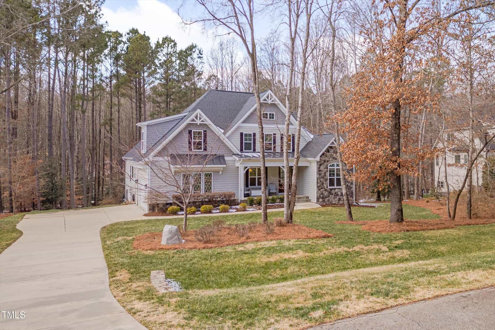 A two-story house with gray siding and stone accents is surrounded by leafless trees. The front yard features a manicured lawn, shrubs, and a concrete driveway. A wooded area is visible in the background under a partly cloudy sky.