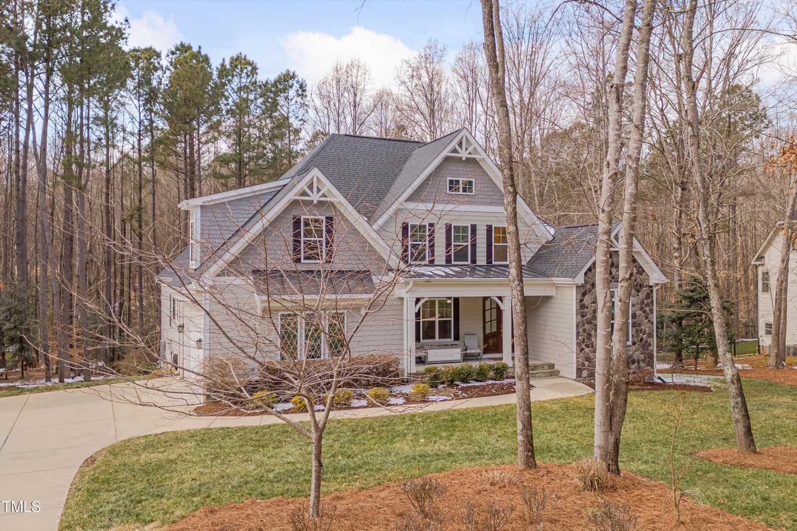 A two-story house with a gray facade and white trim is surrounded by trees. It features a covered front porch, gable roofs, and a neatly manicured lawn. A driveway leads to the side of the house, and there's a wooded area in the background.