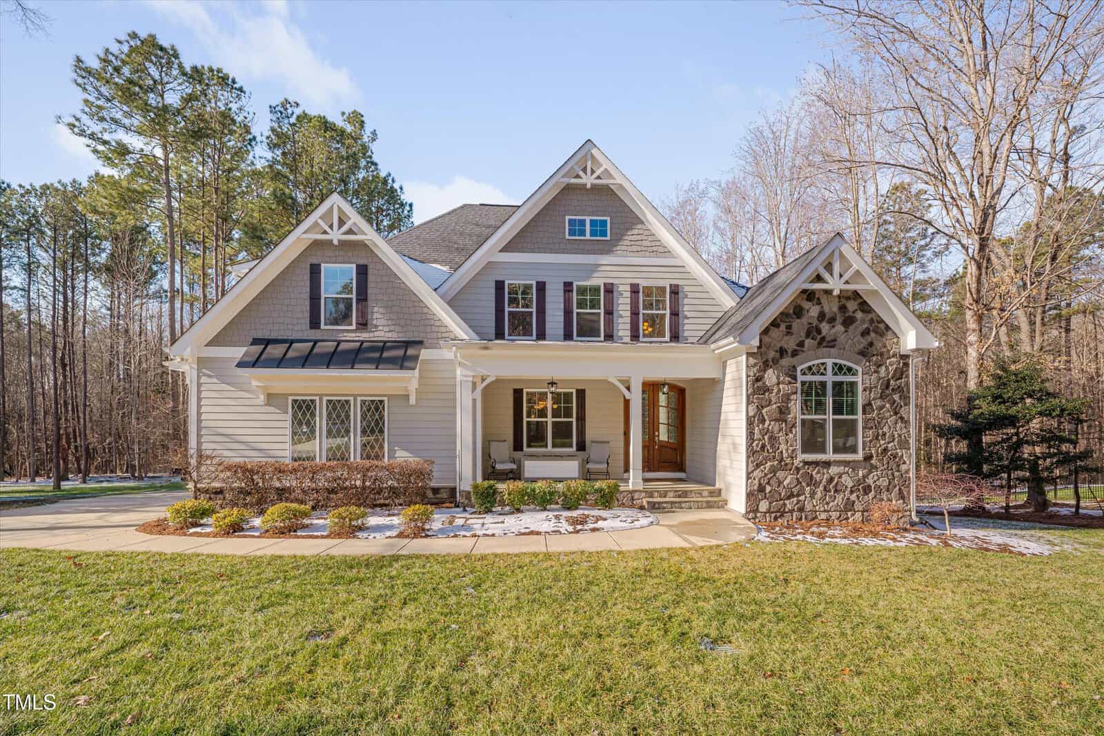 Nestled at 659 Willard Drive, this two-story house in Creedmoor boasts beige siding and stone accents. It features a covered porch with elegant white columns and a dark wooden front door. The peaked roof with dormer windows complements the surrounding trees beneath a clear blue sky.
