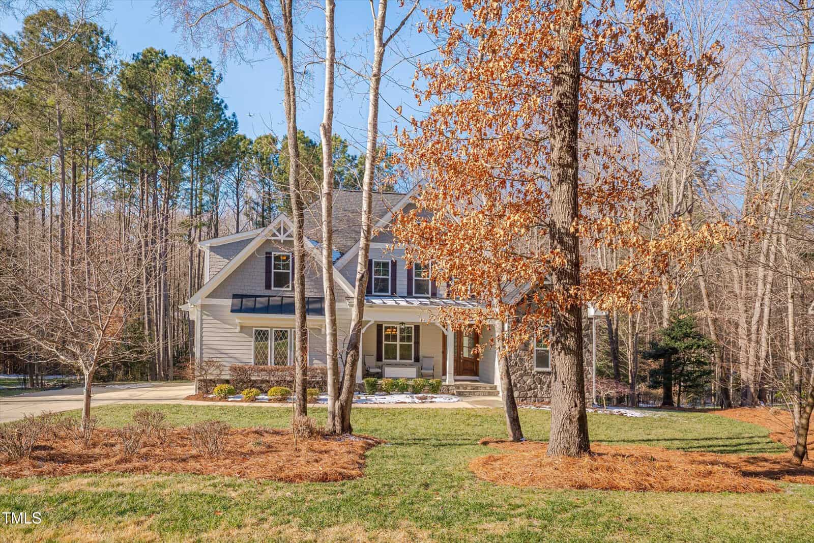 A two-story house with a stone and beige facade is surrounded by trees. There is a manicured lawn and a driveway on the left. A tree with brown leaves is in front, and there are evergreen trees in the background under a clear blue sky.