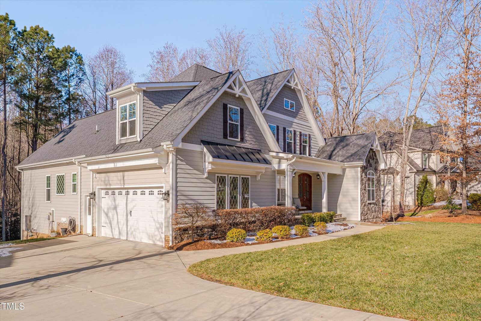 A two-story house with light gray siding and a dark gray roof sits on a curved driveway. The front features a wooden door, white-framed windows, and a two-car garage. Leafless trees and a grassy lawn surround the property.