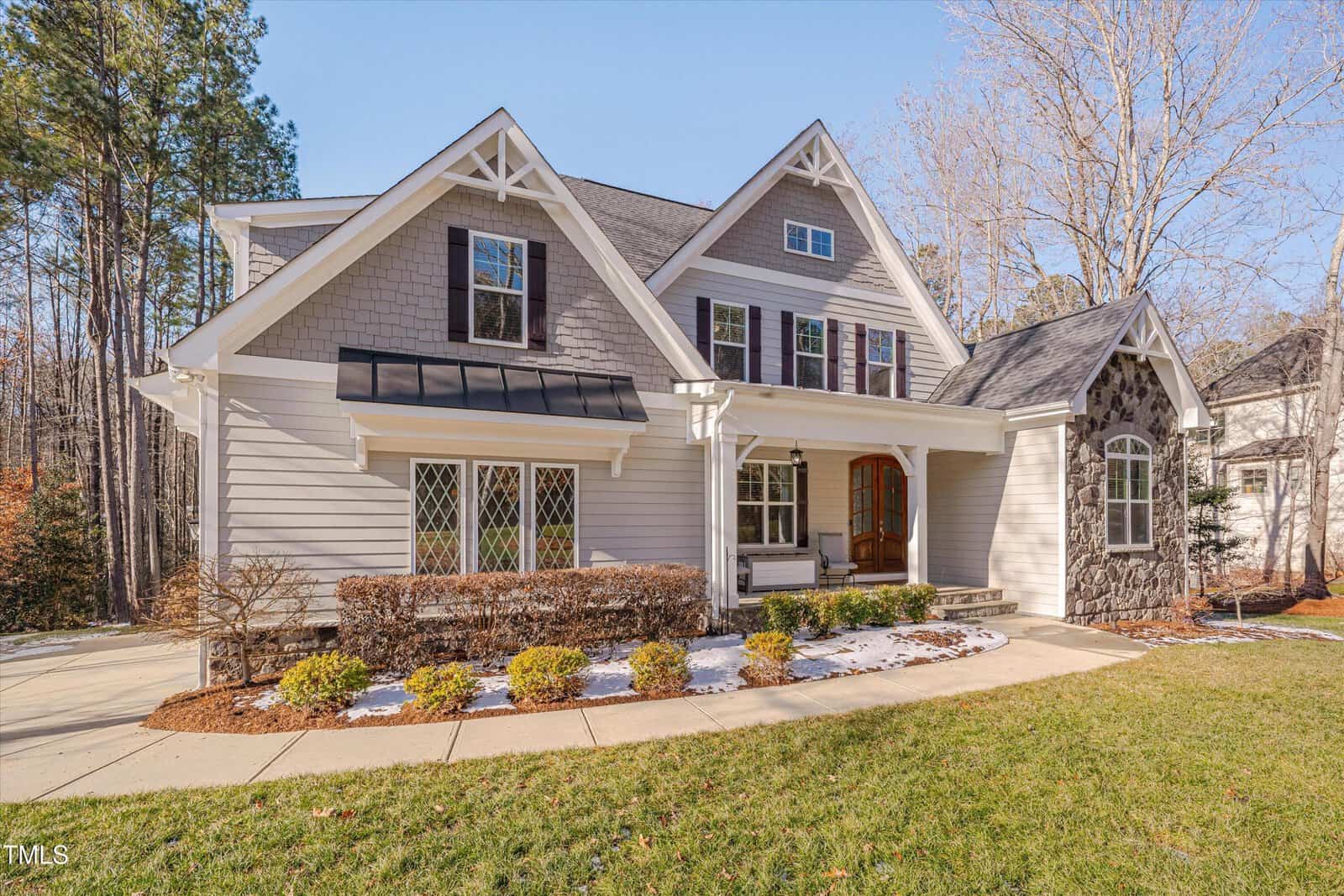 A two-story house with a gray exterior, white trim, and a mix of shingles and stonework. It features peaked roofs, a small porch with columns, a manicured lawn, and a concrete walkway. Trees are visible in the background.