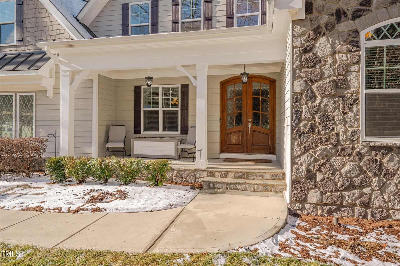 A house with a stone and white siding exterior features a covered front porch with a wooden double door, two chairs, and a bench. Snow and patches of grass are visible on the lawn and along the concrete walkway.