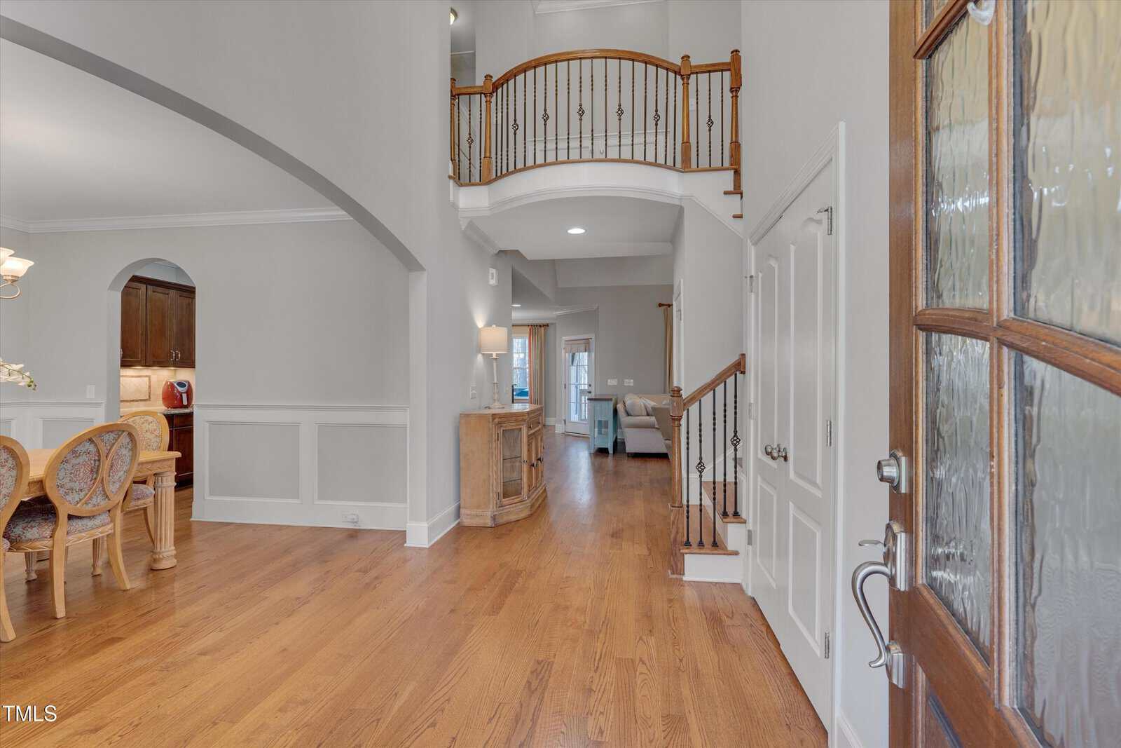 Spacious foyer with wooden floor, leading to a living area. A staircase with wooden railing ascends to a second-floor landing. Dining area with a table and chairs is visible to the left. There's a wooden console and the entrance door has glass panes.