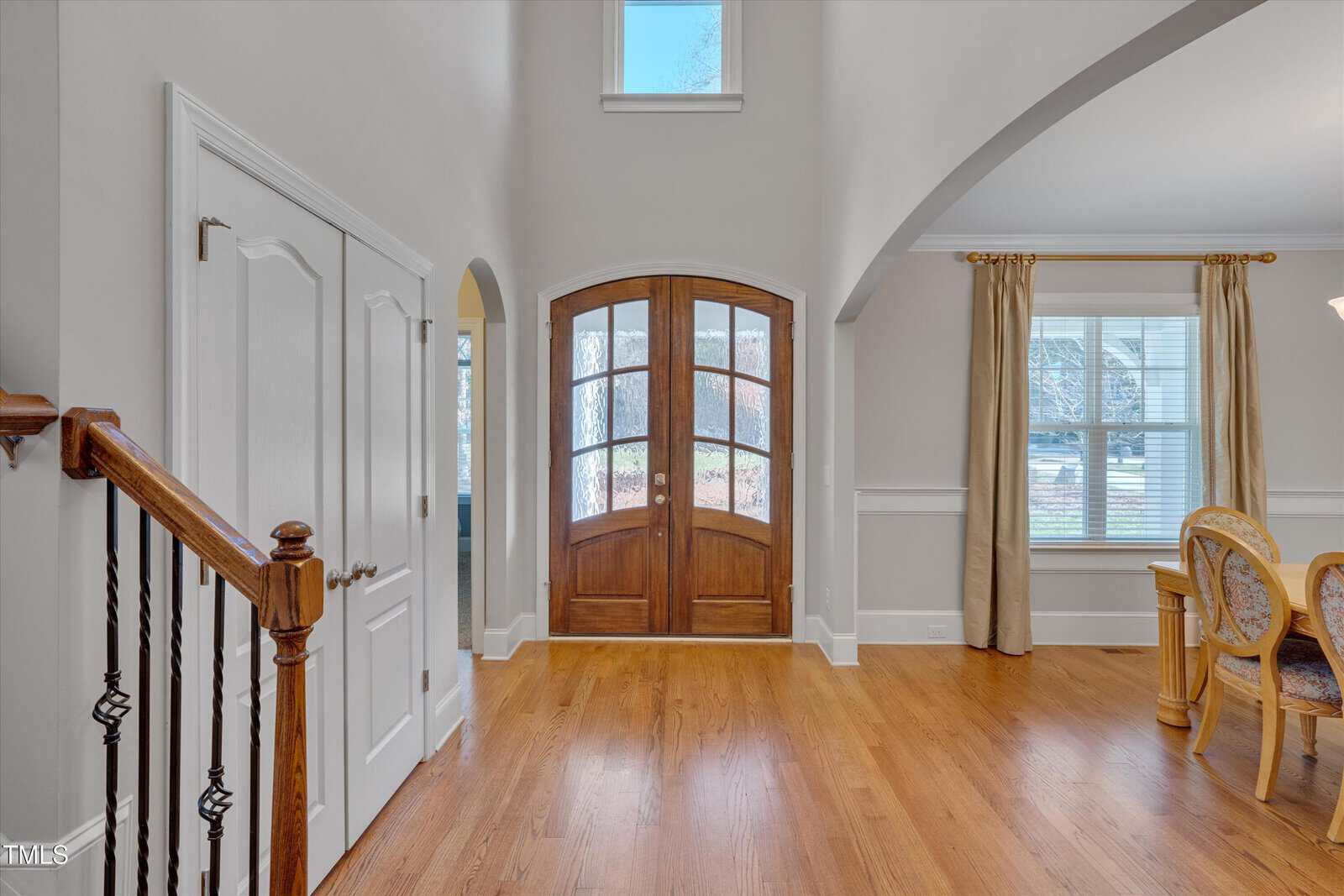 Bright entryway with wooden double doors and arched window above. Adjacent to a dining area with a window and curtains. Hardwood flooring throughout, with a staircase visible to the left and interior doors on the opposite side.