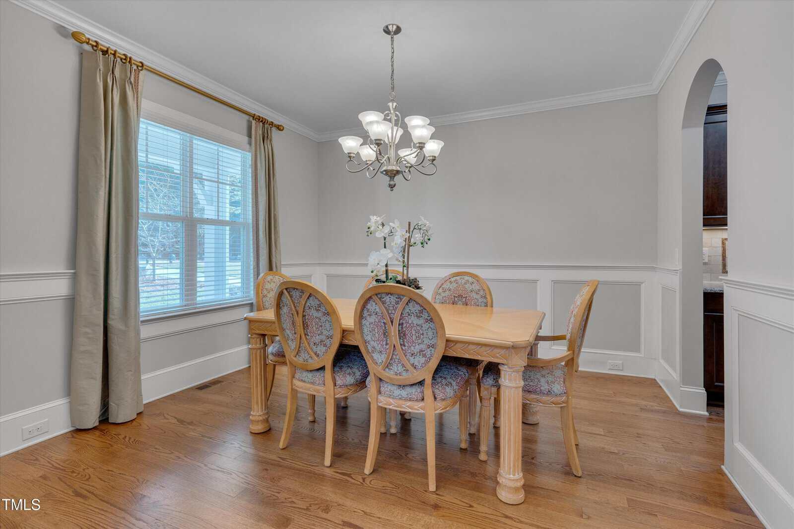 Dining room with light wood floor. Features a wooden table and six upholstered chairs. A chandelier hangs overhead. A window with beige curtains is on the left, and there is an arched doorway leading to another room on the right.