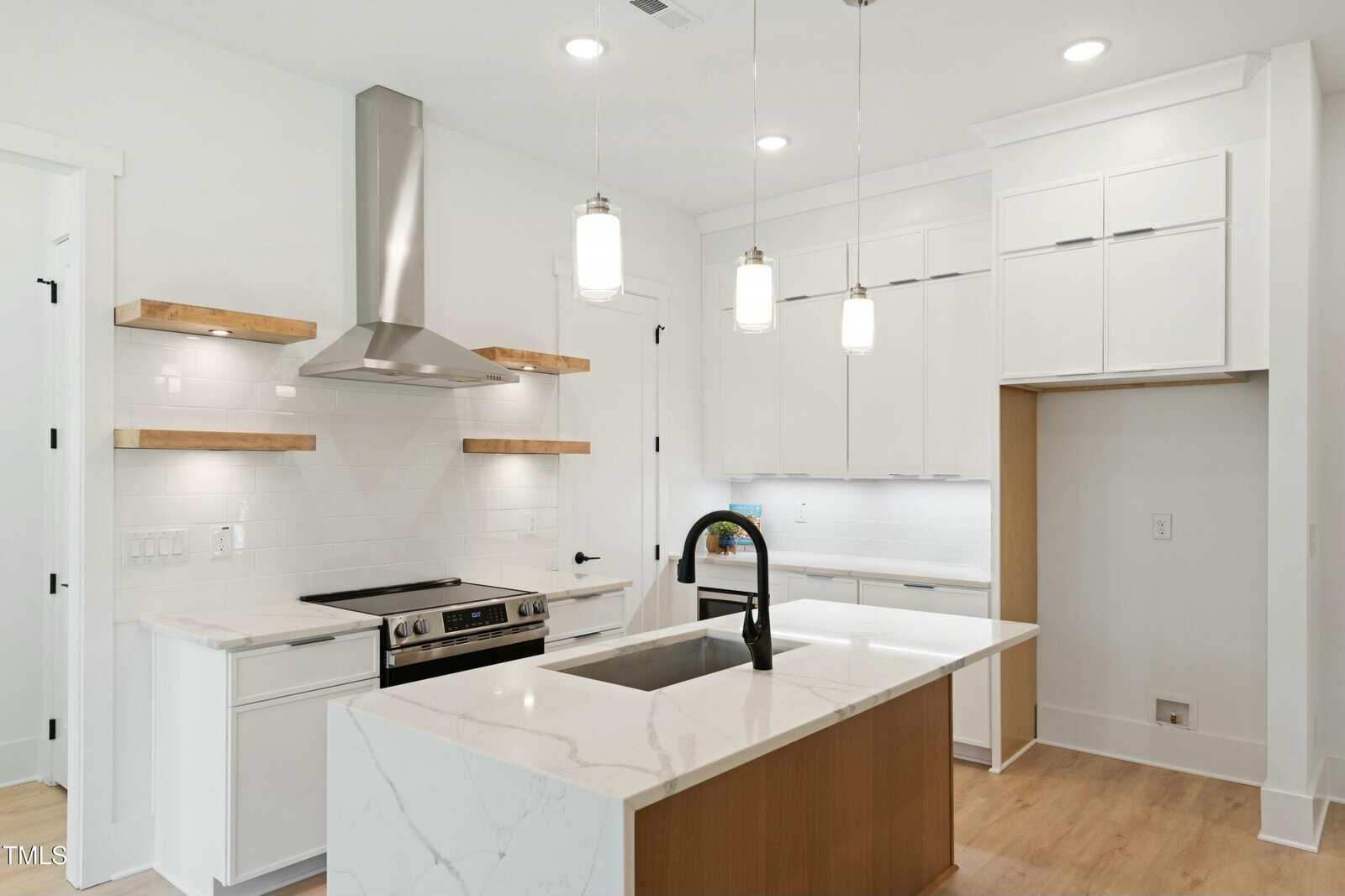 Modern kitchen with white cabinets, a stainless steel range and hood, built-in oven, white subway tile backsplash, wood floating shelves, a large island with a sink and black faucet, and pendant lights overhead.